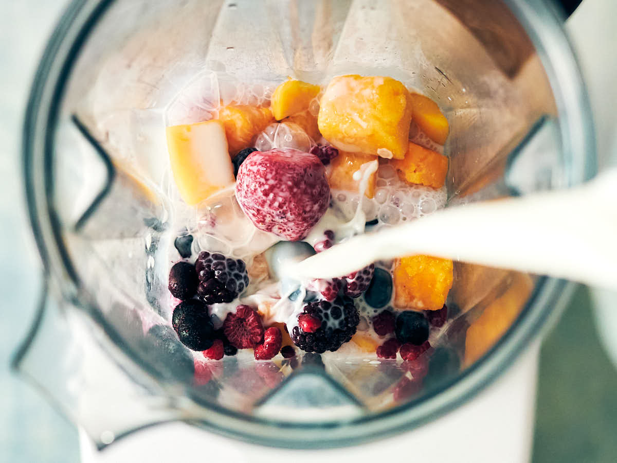 Pouring milk into a blender with frozen mango and berries.