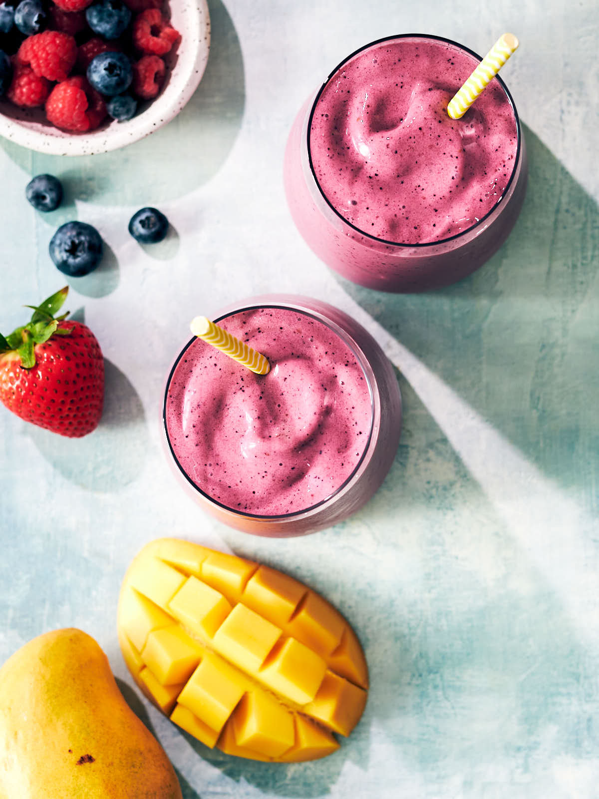Two mango berry smoothies in glasses with straws and fresh fruit beside them.