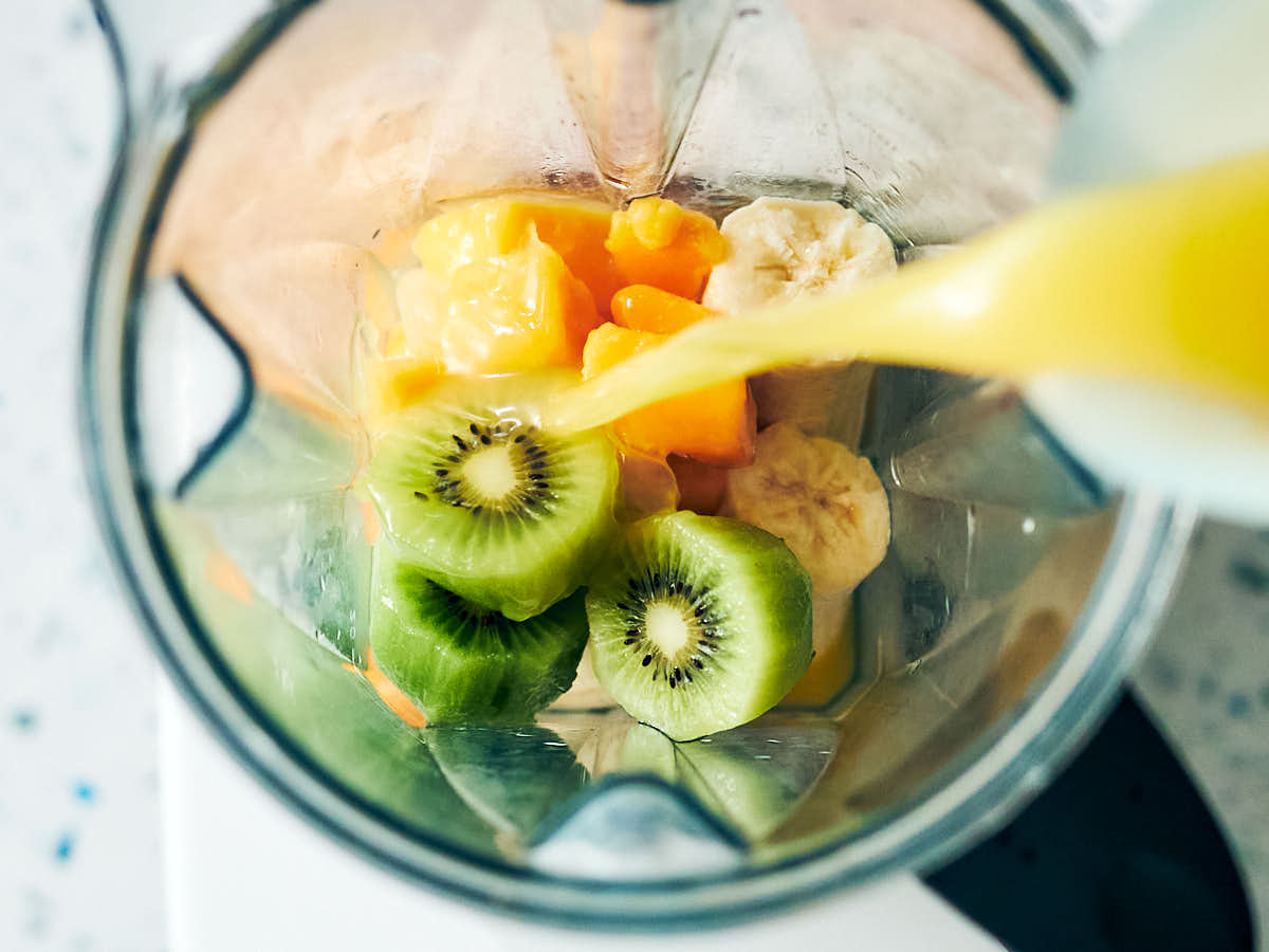 Orange juice being poured into a blender with kiwi, mango, and banana. 