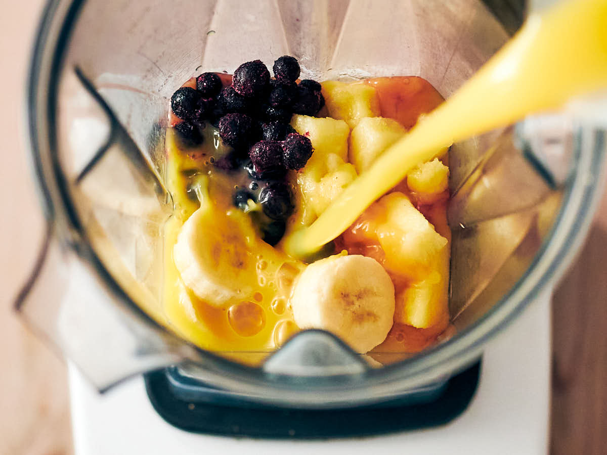 Pouring orange juice into a Vitamix blender with banana, blueberries, and pineapple.