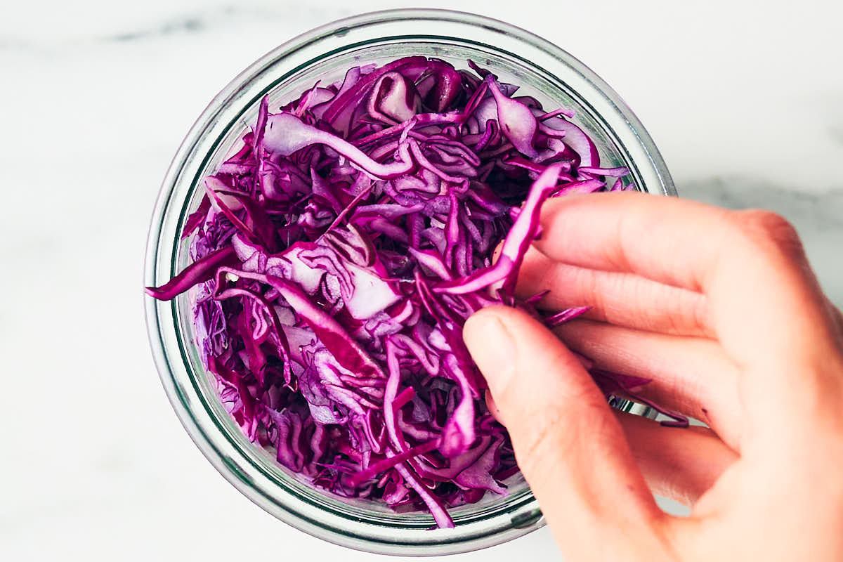 A hand adding red cabbage into a glass jar for pickled cabbage recipe.