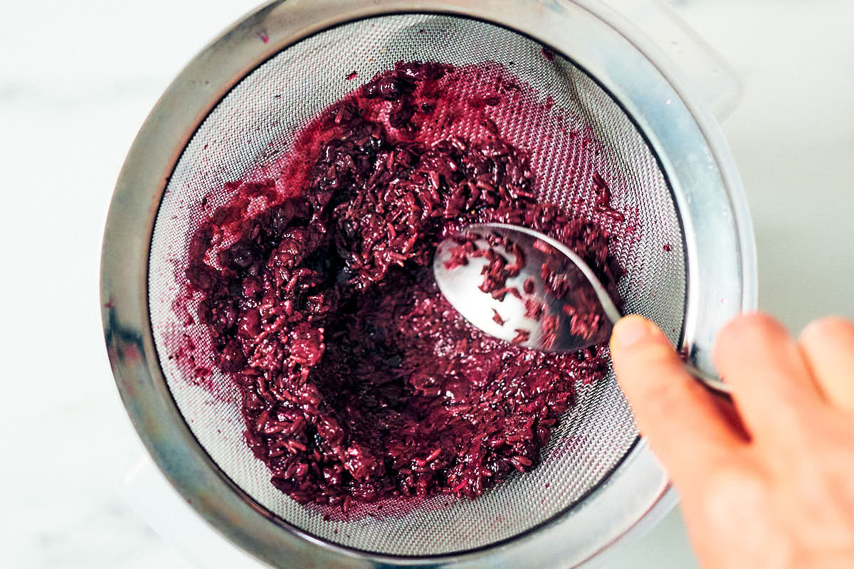 A spoon mashing blueberries into a strainer to extract juice.