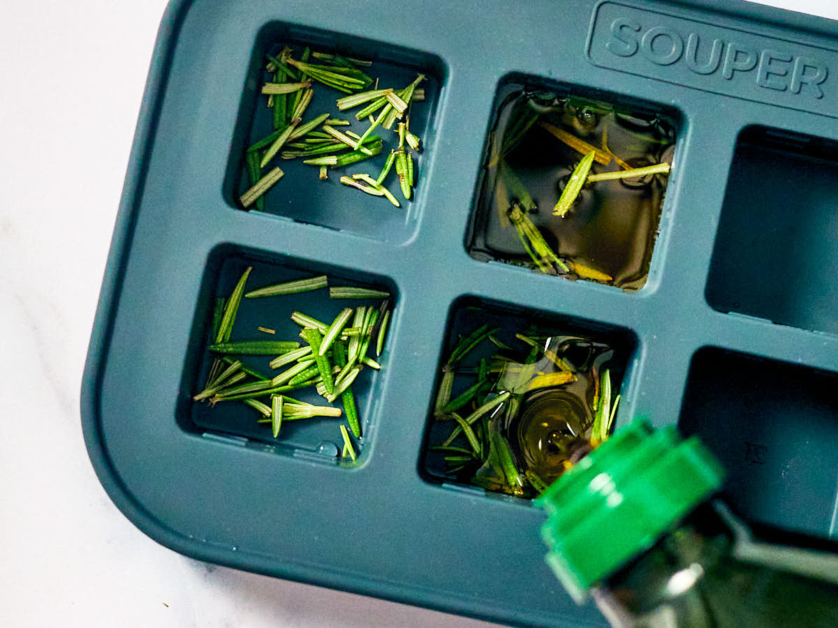 Olive oil being poured into an ice cube tray with fresh rosemary.