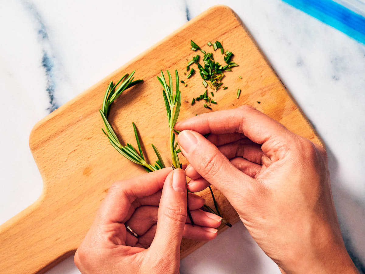 Rosemary leaves being picked off the stem and chopped on a cutting board.