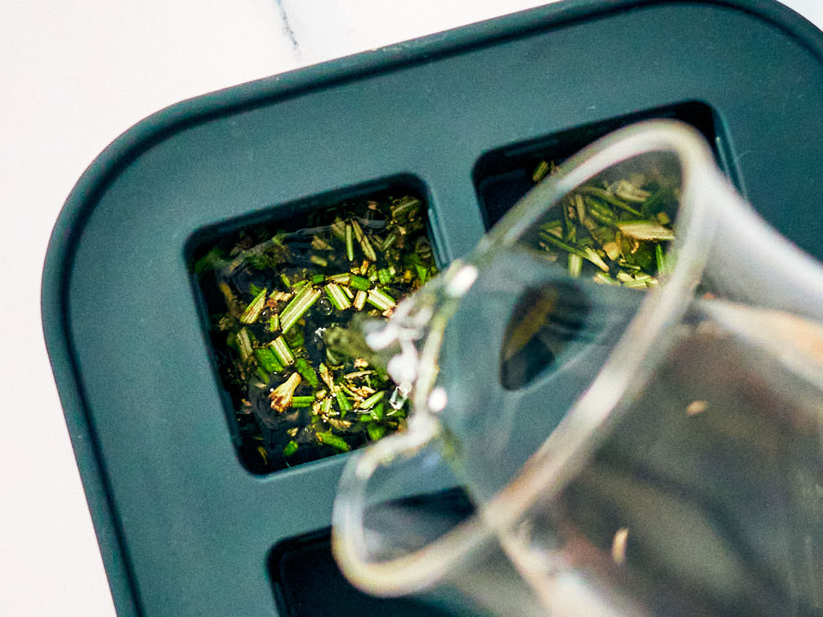 Chopped fresh rosemary in ice cube trays with water being poured on top.