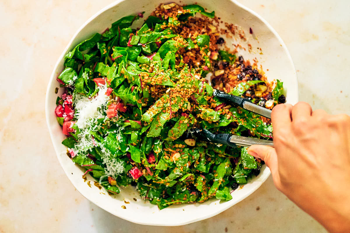 Tongs tossing swiss chard salad with dressing and toppings in a bowl.