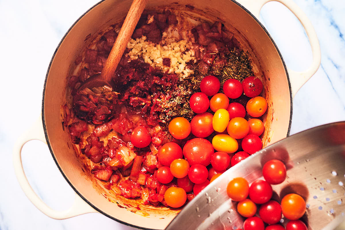 Cherry tomatoes being added to a pot with sundried tomatoes, onion, and garlic.