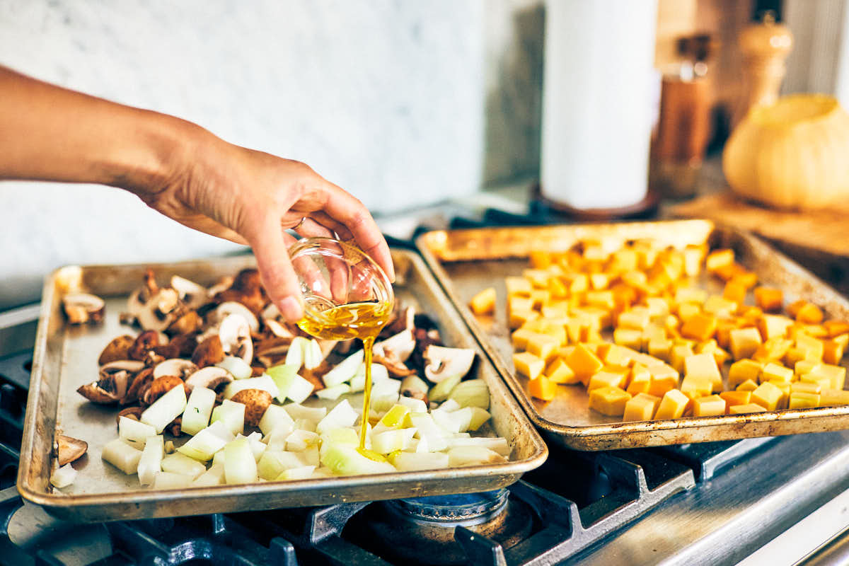 Oil drizzling over butternut squash, mushrooms, and onion on baking sheets.