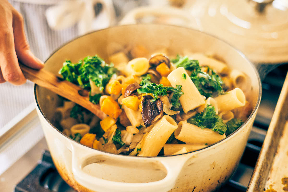 Roasted butternut squash, mushrooms, and kale being stirred into a pot of pasta.