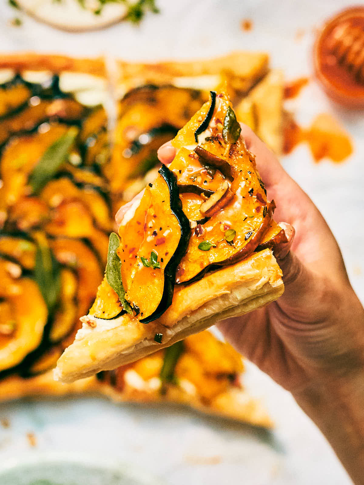 A hand holding a slice of roasted squash tart with feta and honey.