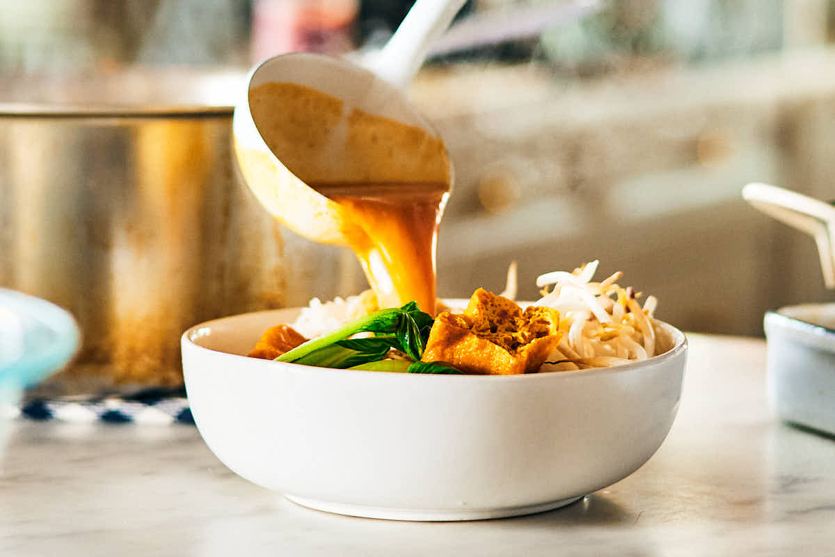 Vegan laksa soup broth being ladled over a bowl of noodles, tofu, and toppings.