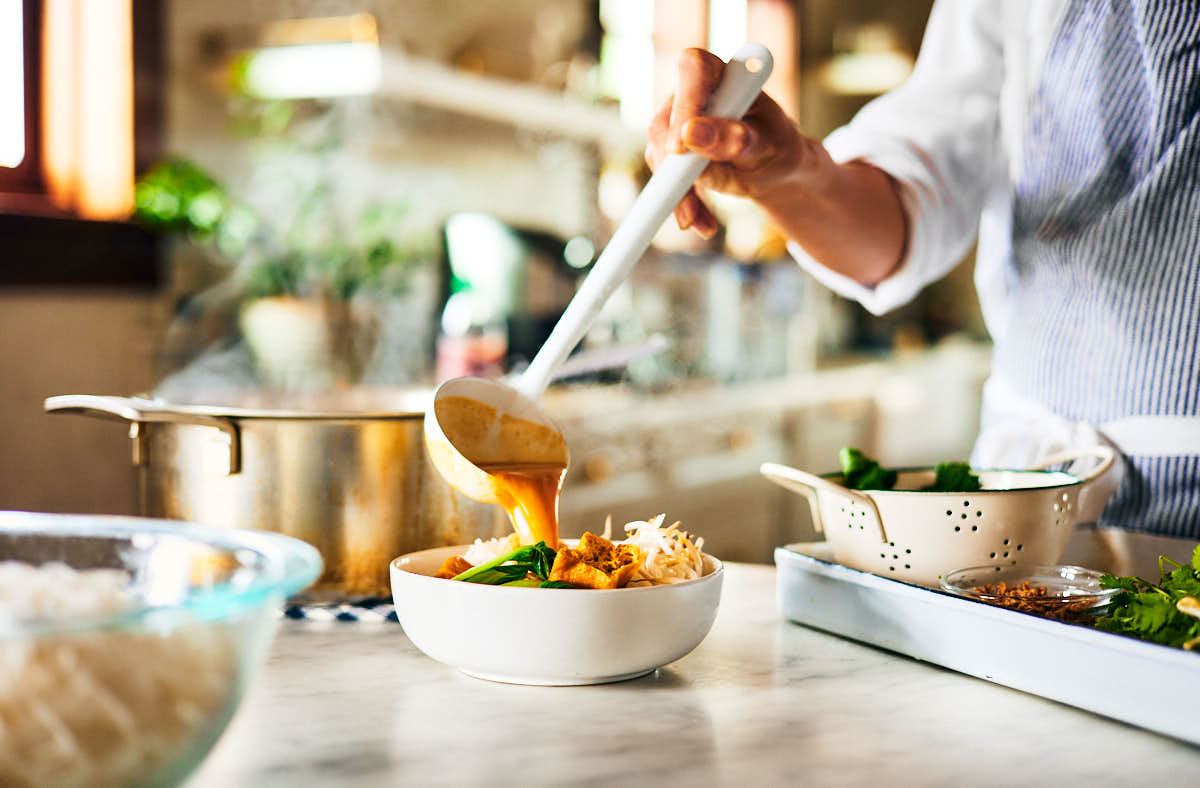 Vegan laksa soup broth being poured over noodles in a bowl.