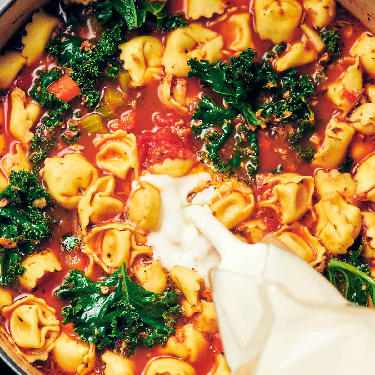 Cream being poured into a pot of Vegetarian Tortellini soup with kale.