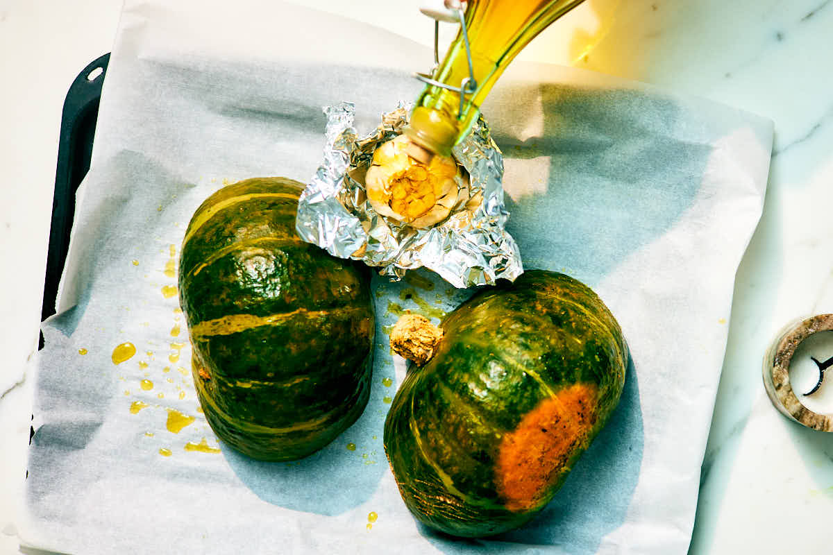 Olive oil being drizzled over garlic and kabocha squash on a baking sheet.