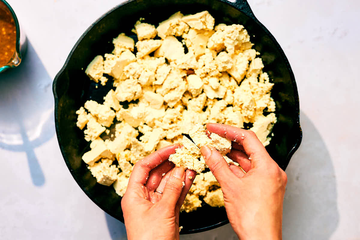 Hands crumbling tofu into a hot skillet to be pan fried.