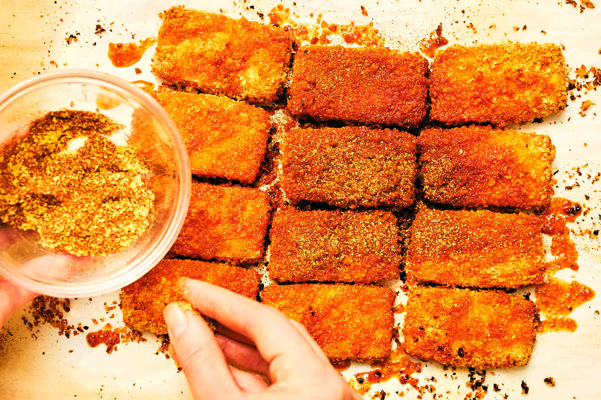 Spices being sprinkled over crispy hot tofu for a Nashville-style vegan sandwich.