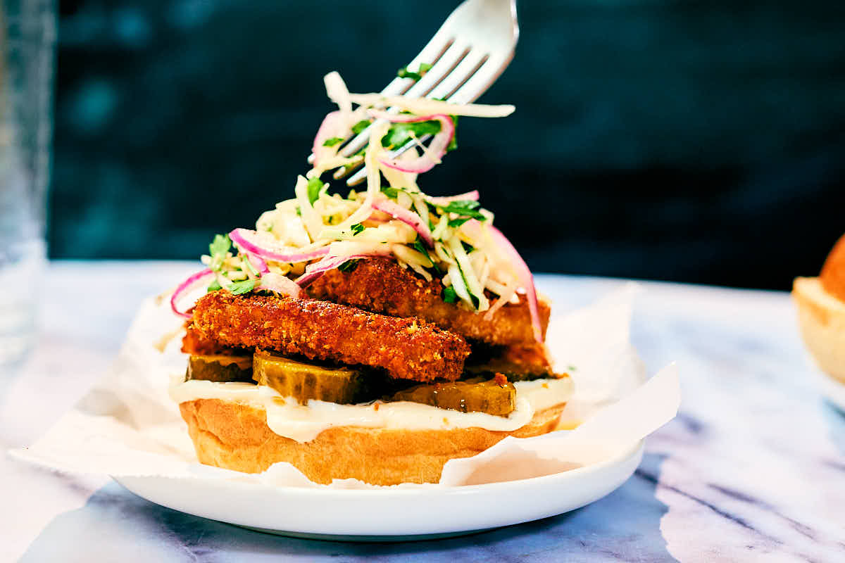 Slaw being added on top of a Nashville Hot Tofu Sandwich on a plate.