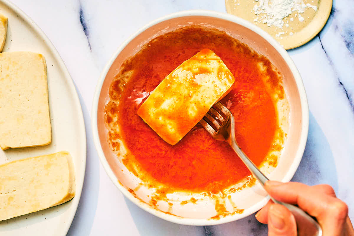 Slices of tofu being dipped into a spicy sauce for Nashville Hot Tofu.