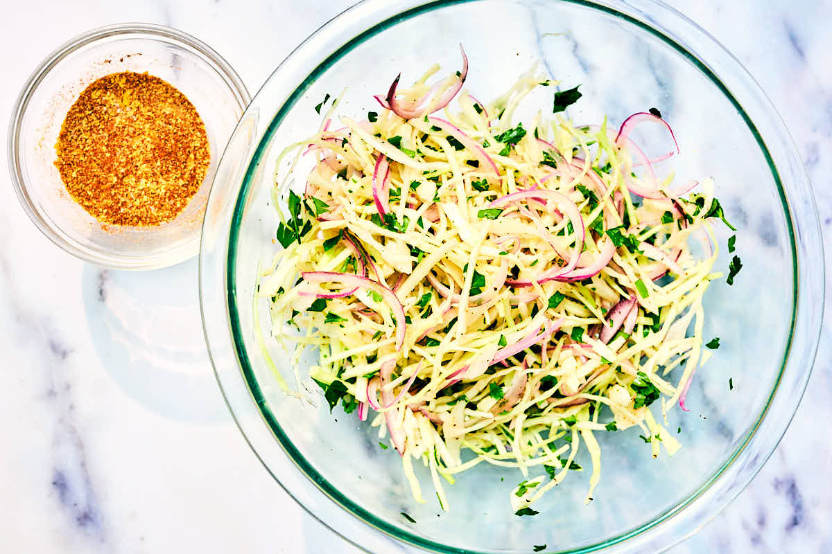 A bowl of green cabbage coleslaw and a bowl of spices on a marble counter.