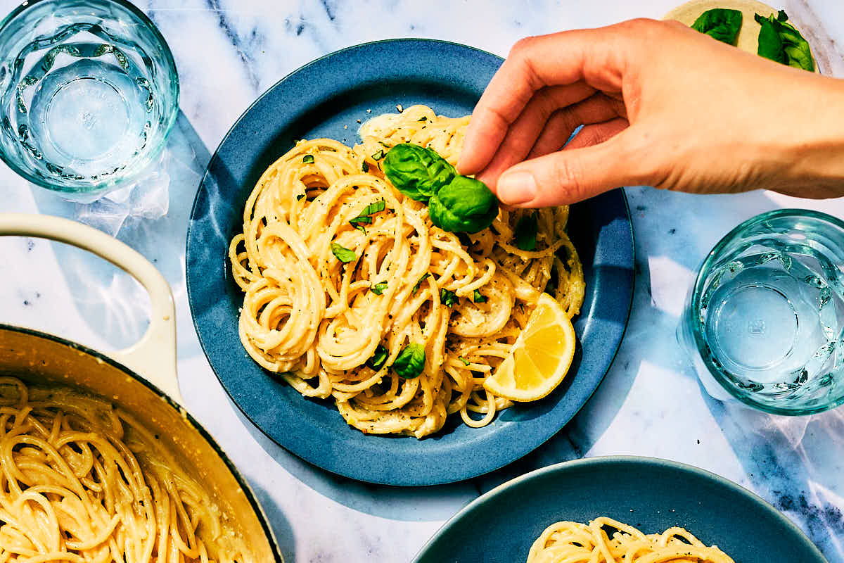 Fresh basil being placed on top of a plate of lemon pesto pasta.