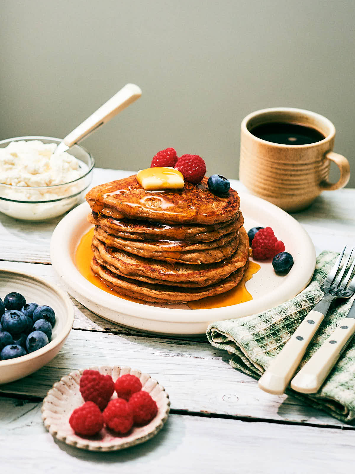 A stack of high protein Cottage Cheese pancakes on a plate with berries, maple syrup and butter.